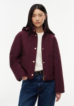 Unge kvinde iført en burgunder quiltet jakke med gyldne knapper, hvid sweater og blå jeans, stående med den ene hånd i lommen.