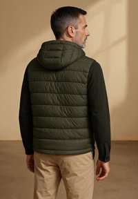Gevoerde capuchonvest in donkergroen, met een soepele textuur en een getailleerde pasvorm, gecombineerd met lange zwarte mouwen en khaki broek.