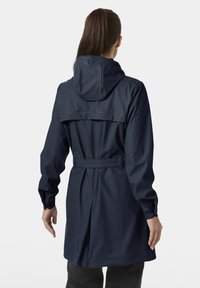 Vrouw met lang bruin haar die een marineblauwe regenjas met een ceintuur en capuchon draagt, gezien vanaf de achterkant tegen een effen lichte achtergrond.