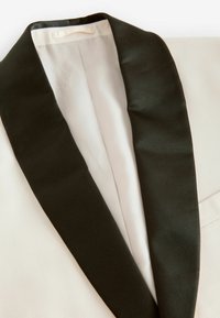 Veste de smoking blanche avec un col châle en satin noir. L'intérieur présente un tissu léger et lisse avec une texture subtile.