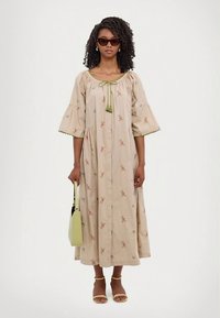 Beige cotton dress with floral embroidery, puff sleeves, and a tie detail at the neckline. Paired with yellow sandals and a green handbag.