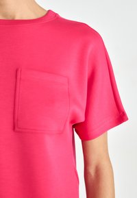 T-shirt rose vif à manches courtes en tissu lisse, avec une poche sur le devant et un col rond. Détails de couture soignés.