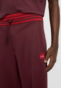Burgundy joggers with a red striped elastic waistband, drawstring closure, and embroidered logo featuring a bull and initials on the side.
