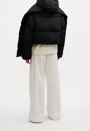Personne portant une doudoune noire courte par-dessus un pull blanc et un pantalon blanc à jambes larges, debout devant un fond clair uni.
