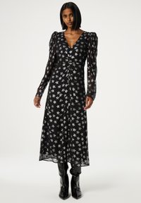 Schwarzes Kleid mit silbernem Sternenmuster, langen durchsichtigen Ärmeln, V-Ausschnitt, geraffter Taille und fließender Silhouette, kombiniert mit schwarzen, glänzenden Stiefeln.