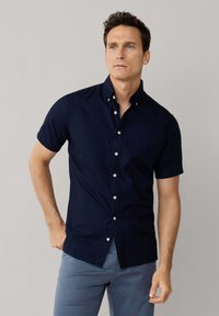 Muž v námořnicky modré krátkosrsté košili s knoflíčky a světle modrých kalhotách, stojící s jednou rukou v kapse na jednobarevném pozadí.