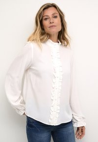 White blouse with ruffled front and button closure, long sleeves with gathered cuffs, made of lightweight fabric, paired with denim jeans.