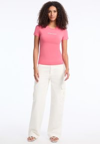 Rožinė prigludusi marškinėliai su "GUESS JEANS" logotipu, derinami su smėlio spalvos plačiomis krovinių kelnėmis, turinčiomis šonines kišenes. Visoje medžiagoje lygi tekstūra.