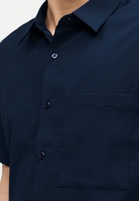 Άνδρας που φοράει σκούρο ναυτικό κοντομάνικο πουκάμισο με υφή και κουμπιά, με τσέπη στο στήθος που διαθέτει διακριτικό λογότυπο CK.