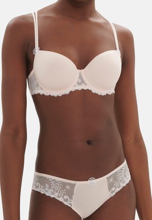 Kobieta ubrana w dopasowany bladoróżowy biustonosz i majtki z przezroczystymi koronkowymi detalami i kwiatową haftem na jednolitym tle.