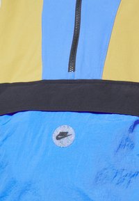 Flerfarget Nike-jakke med glidelås foran, med blå, gul og svart paneler, og et sirkelformet logo-patch på nederste front.