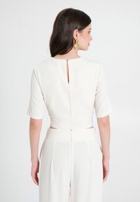 Completo bianco a due pezzi con top corto caratterizzato da un dettaglio di bottoni sulla schiena e pantaloni a gamba larga con chiusura a cerniera e pieghe.