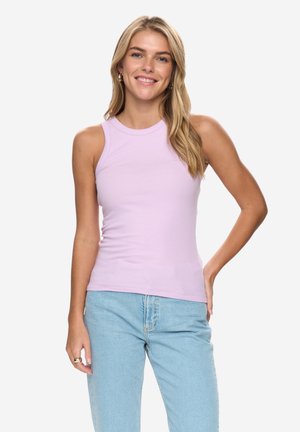 Femme souriante portant un haut sans manches violet clair et un jean bleu clair, debout avec une main sur la hanche devant un fond uni.