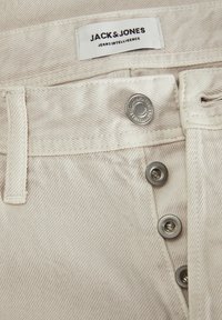 Bež traperice s vidljivom oznakom "JACK & JONES JEANS INTELLIGENCE", s srebrnim gumbom i suptilnom teksturom riblje kosti.