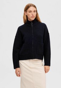 Navyblauwe gebreide ritsjack met een hoge kraag, twee voorzakken en een cropped pasvorm. Zachte structuur en een ontspannen silhouet.