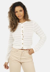 Biały sweter z dzianiny o teksturowanym wzorze, z złotymi guzikami z przodu i prążkowanymi mankietami. Noszony z beżowymi spodniami z wysokim stanem.