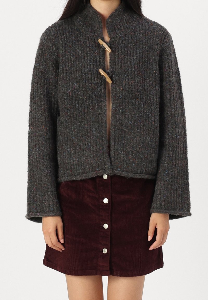 Femme portant un cardigan en grosse maille gris foncé avec des fermetures à bascule en bois, sur une jupe en velours côtelé bordeaux à boutons sur le devant, debout devant un fond blanc.