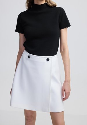 Kobieta ubrana w czarny, krótki top z półgolfem oraz białą spódnicę w linii A z dwoma czarnymi guzikami w talii.