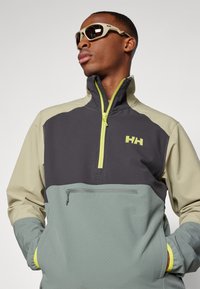 Hochschlupfjacke in Grau, Beige und Grün. Aus strukturiertem Stoff, mit kontrastierenden Reißverschlüssen und einem Logo auf der Brust. Vordertasche mit Reißverschluss.