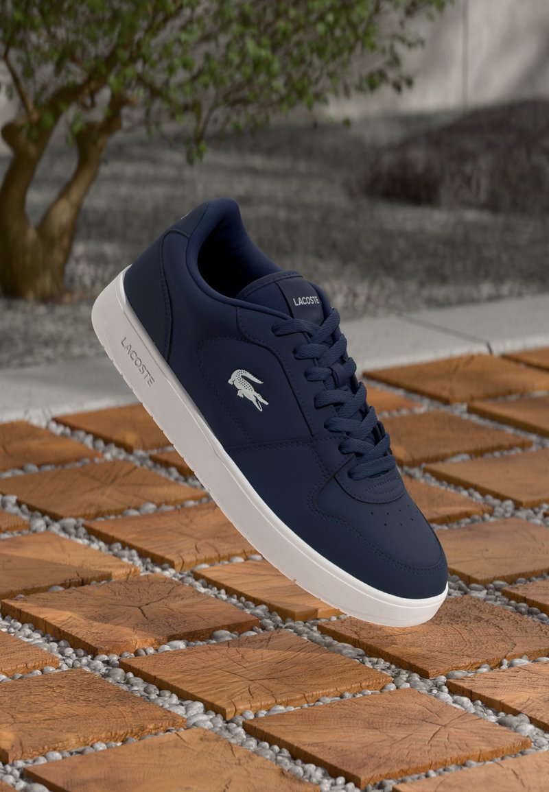 Baskets Lacoste bleu marine avec semelle et logo blancs, flottant au-dessus de carreaux en bois avec des interstices en gravier, fond d'arbres et de pierres.