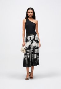 Μαύρο τοπ με έναν ώμο, συνδυασμένο με μίνι φούστα πλισέ σε μαύρο και άσπρο, με λουλουδάτο σχέδιο. Το outfit περιλαμβάνει μαύρες ψηλές γόβες με λουριά και μπεζ τσάντα.