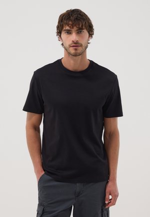 Mann mit kurzen lockigen Haaren, der ein schwarzes T-Shirt und dunkle Cargohosen trägt, steht vor einem schlichten hellen Hintergrund.