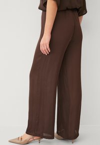 Brune brede bukser i let stof, med en afslappet pasform, elastisk talje og en subtil tekstur. Sammen med beige højhælede sko.