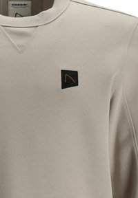 Světlé šedé tričko s texturovanou látkou, žebrovaným výstřihem a černou hranatou nášivkou s oranžovým motivem na levé straně hrudi.