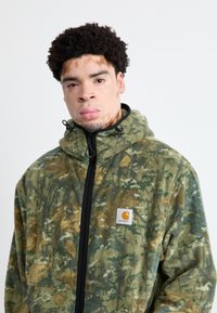 Jachetă din fleece cu model camuflaj verde și brown, cu fermoar și guler înalt, având un fermoar negru și o petic albă cu logo pe pieptul stâng.