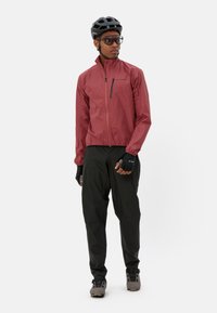 Rote leichte Jacke mit Reißverschlusstasche, kombiniert mit schwarzen Hosen und Handschuhen. Zubehör umfasst einen schwarzen Helm und eine Sonnenbrille.