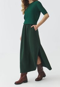Groene geribbelde gebreide top gecombineerd met een lange, vloeiende groene rok met zijsplitten, aangevuld met een paar bruine enkellaarzen.
