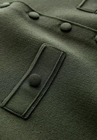 Mørkegrønt stof med en tekstureret finish, med en klaplomme, tre knapper og pænt syede kanter.