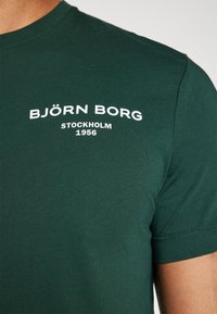 Mørkegrønn bomulls t-skjorte med ribbet rund hals, med hvit tekst "BJÖRN BORG STOCKHOLM 1956" på forsiden. Korte ermer.