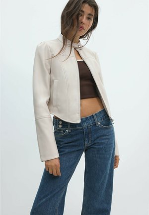 Frau trägt eine hellbeige, kurz geschnittene Jacke über einem braunen Crop-Top, kombiniert mit hoch taillierten blauen Jeans vor einem schlichten hellen Hintergrund.