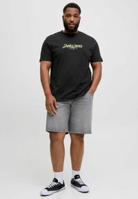 Fekete grafikás póló, arany betűkkel "Jack & Jones" felirattal, szürke farmernadrággal és fekete sportcipővel, fehér talppal és zoknival párosítva.