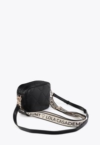 Svart quiltad crossbody-väska med ett guldspänne och en beige rem med texten "LOLA CASADEMUNT". Mot en enfärgad vit bakgrund.