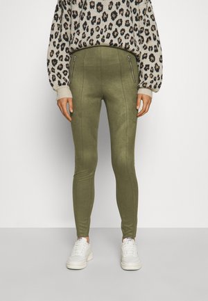 Person trägt hoch taillierte olivgrüne Leggings mit Frontreißverschlusstaschen, Leopardenmuster-Pullover und weiße Turnschuhe und steht vor einfarbigem Hintergrund.