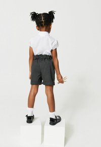 Pantaloni corti grigi con vita arricciata, risvolti e una camicia bianca a maniche corte. Scarpe nere con calze di pizzo e materiali artistici in mano.