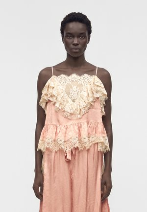 Γυναίκα με σκούρο δέρμα φοράει ροζ σατέν φόρεμα με μπεζ δαντελωτά βολάν και λεπτούς τιράντες, στέκεται μπροστά σε λευκό φόντο.