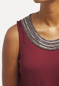 Top senza maniche color borgogna con scollatura a perline texturizzate, caratterizzato da accenti argento e nero in un design circolare a strati.