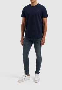 T-shirt en coton bleu marine à col ras du cou et manches courtes, associé à un jean slim et des baskets blanches. Marque minimaliste sur le t-shirt.