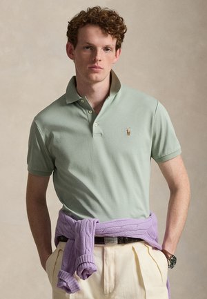 Ung mann med krøllete hår iført en lys grønn polotrøye, kremfargede bukser og en lavendelfarget genser knyttet rundt livet.