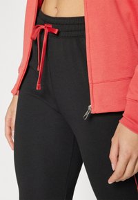 Koralo spalvos zip-up džemperis su raukšlėtu rankogaliu, derinamas su juodomis pasiūtomis kelnėmis, turinčiomis raištelio juosmenį ir užtrauktuku ties kulkšnimi.