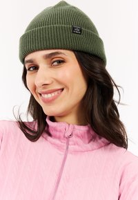 Groene geribbelde gebreide beanie met een gestikt label waarop "PROTEST HOUSE OF SNOW" staat. Draagt een roze jack met rits dat een gestructureerd patroon heeft.