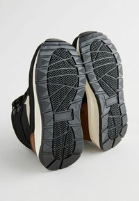 Baskets noires et brunes avec une semelle en caoutchouc texturé, présentant un motif en zigzag, et un haut en tissu. Comprend des détails sur les côtés et des sangles réglables.