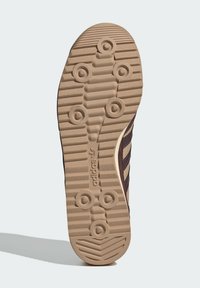 Semelle de sneaker marron avec un motif de grip texturé, présentant des indentations circulaires et des lignes horizontales, accentuée par une bande beige et un logo.