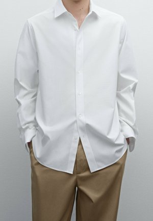 Homme portant une chemise blanche ample à manches longues boutonnée et un pantalon beige, mains dans les poches, devant un fond uni.
