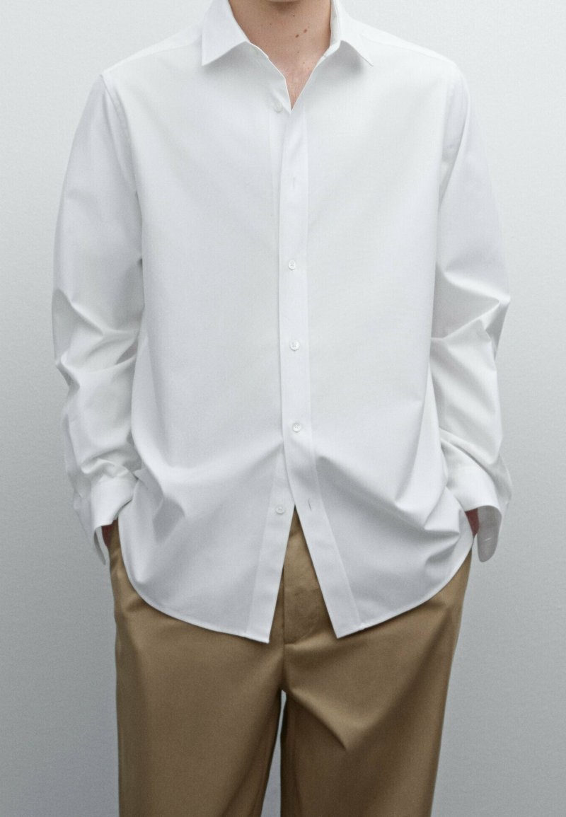 Homme portant une chemise blanche ample à manches longues boutonnée et un pantalon beige, mains dans les poches, devant un fond uni.
