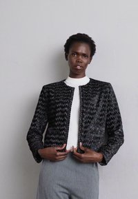Black and silver textured jacket with zigzag pattern, round neckline, and no fastenings, worn over a white top and gray pants.