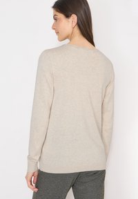 Femme aux longs cheveux bruns portant un pull beige à manches longues et un pantalon gris, vue de dos sur un fond blanc uni.